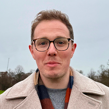Tom is wearing glasses and smiling towards the camera. He is outside and wearing a coat and scarf.